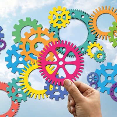 Värikkäitä hammasrattaita, kädessä pinkki hammasratas. Taustana sininen taivas ja poutapilviä