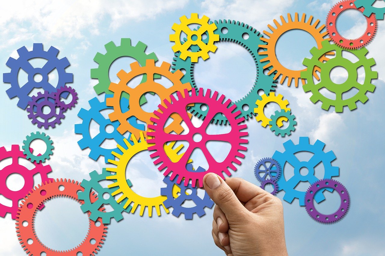 Värikkäitä hammasrattaita, kädessä pinkki hammasratas. Taustana sininen taivas ja poutapilviä