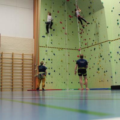 liikuntasalin kiipeilyseinällä kaksi lasta kiipeää seinää ylös, kaksi aikuista varmistaa kiipeäviä lapsia